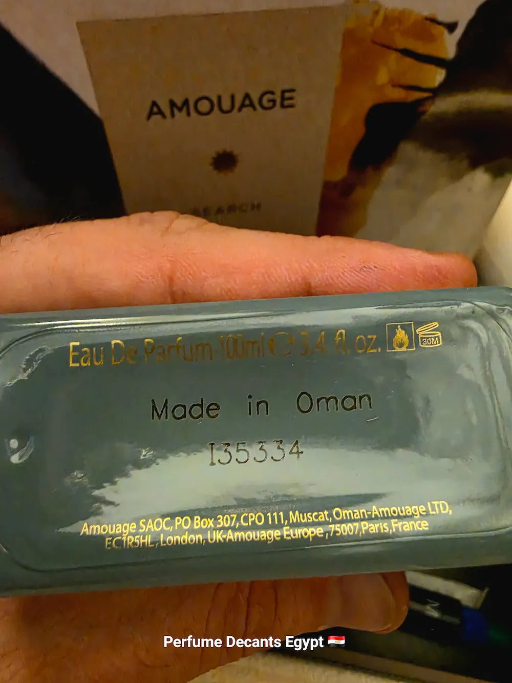 A hand holding a translucent blue-green perfume bottle with gold text, including 'Made in Oman' and 'Eau De Parfum 10ml'. A cardboard box labeled 'AMOUAGE' is visible in the background.