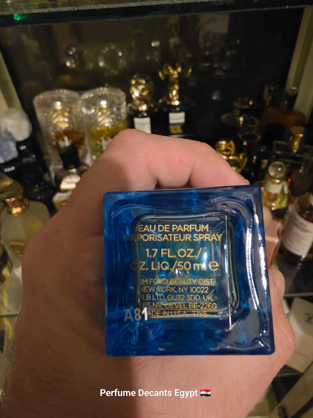 A hand holding a small square blue glass perfume bottle with gold text reading 'EAU DE PARFUM / SPRAY' and volume details, set against a dark shelf background with other perfume bottles.