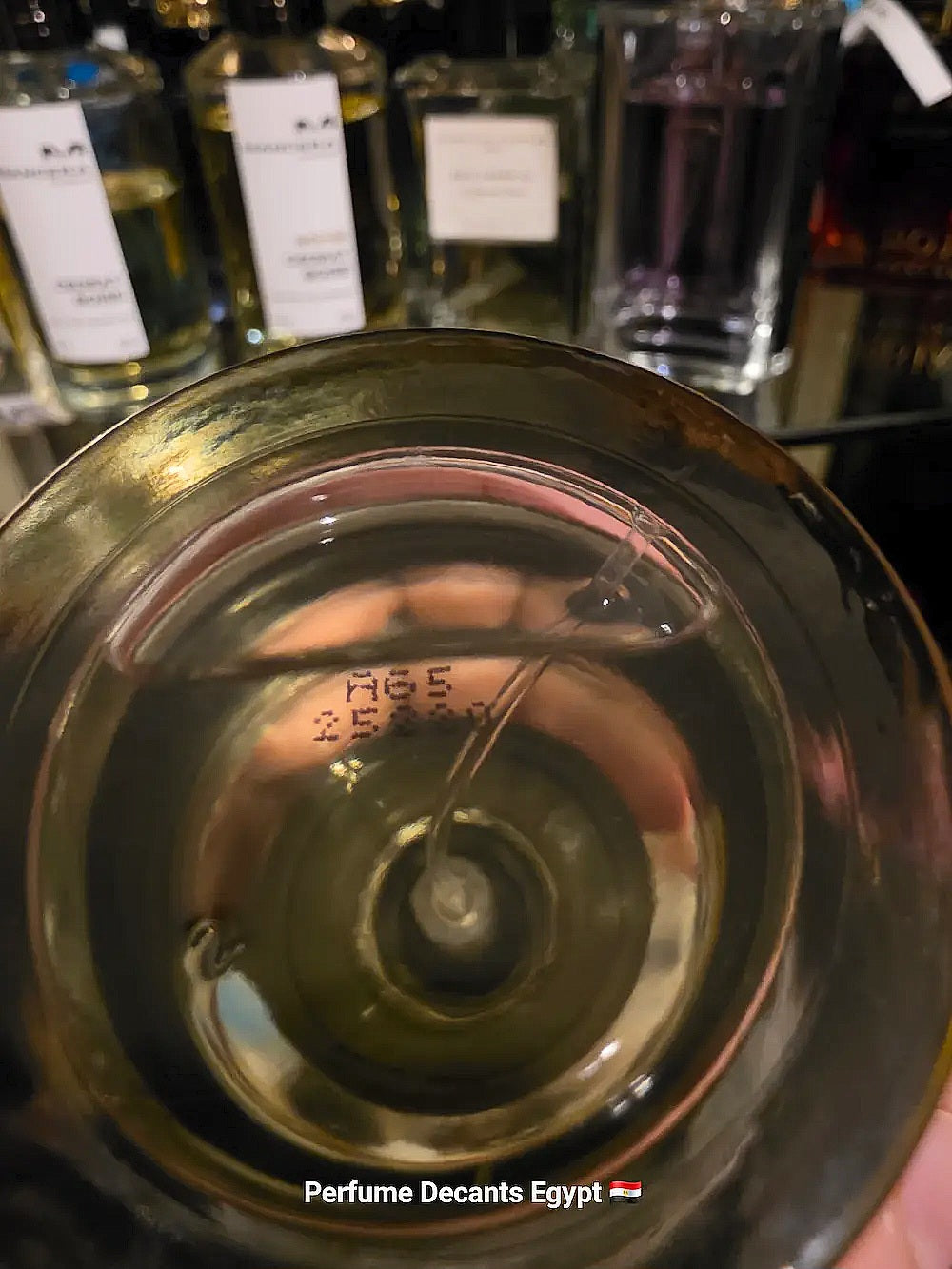 A close-up view from inside a perfume bottle, showing the clear spray nozzle dispensing fragrance. The background is a blurred row of other perfume bottles on a shelf, with a hand holding the bottle at the
Batch number.