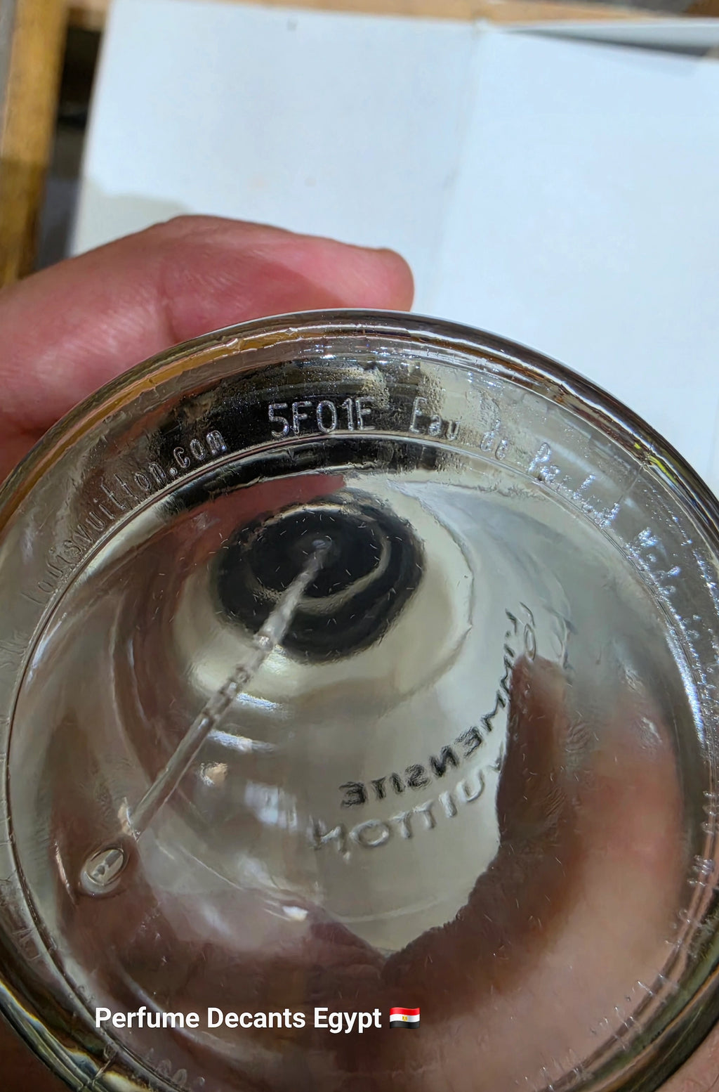 A close-up of a perfume bottle's clear glass bottom, held by a hand. The glass shows embossed text including '5F01F' and 'www.perfumedecantsegypt.com www.louisvuitton.com', with a white atomizer spray wand inserted into the glass.
