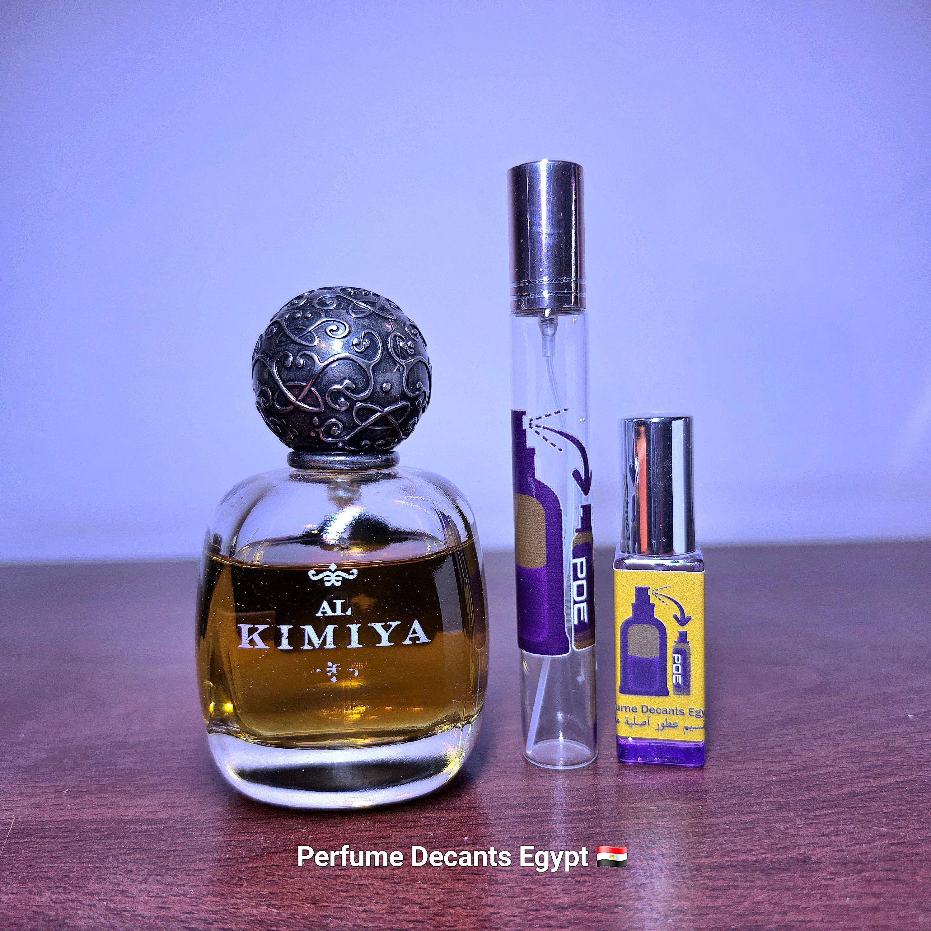 A perfume bottle labeled 'AL KIMIYA' with a dark ornate cap stands next to two travel perfume atomizers, one clear with purple and black labeling and one small clear bottle with a yellow label, all on a wooden surface against a light purple background.