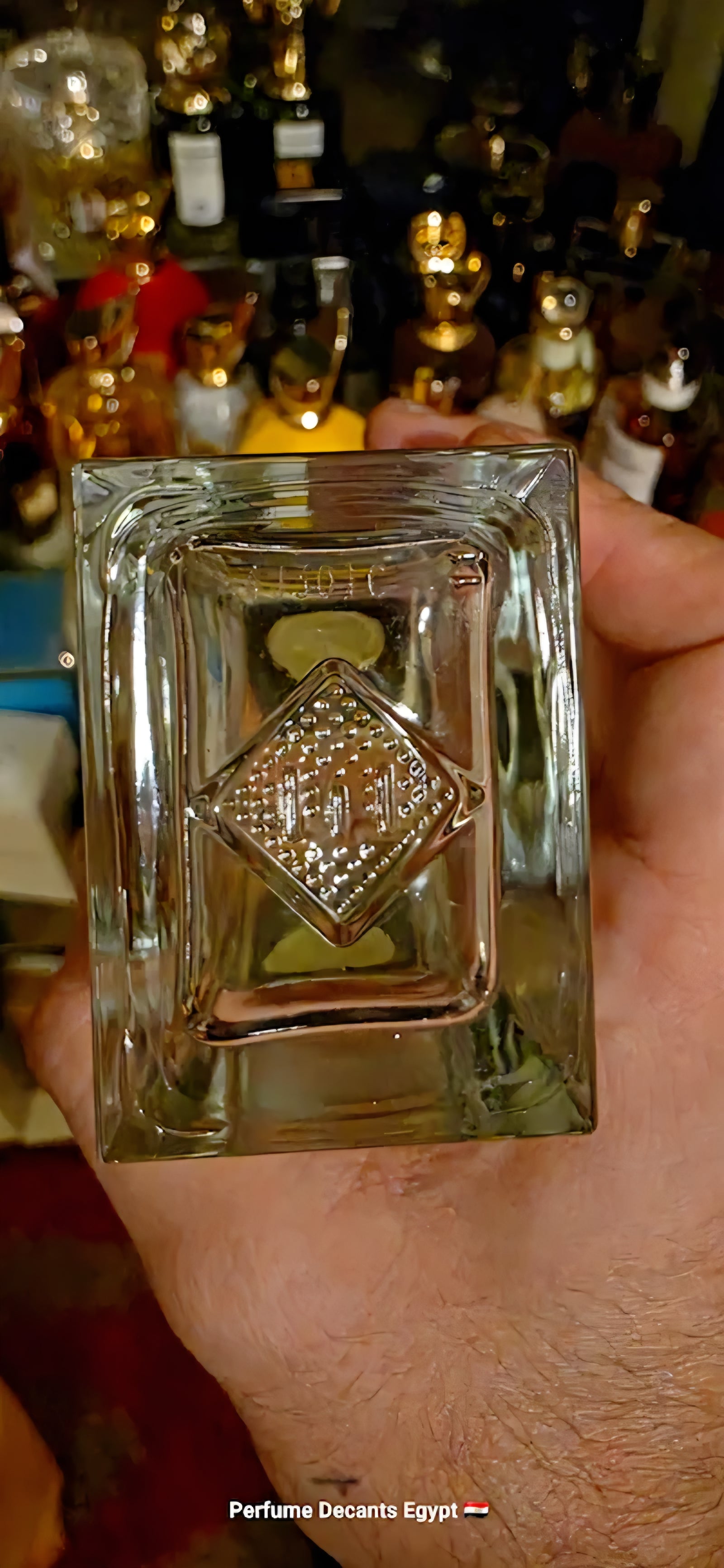 A hand holding a clear square glass old  Batch code perfume bottle with a diamond-shaped embossed logo in the center, set against a blurred background of many perfume bottles.